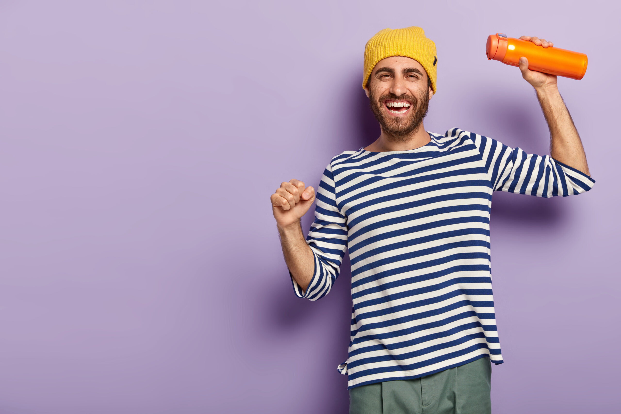 Photo of energized cheerful man tourist dances playfully, dressed in fashionable clothes, carries thermos with aromatic drink, smiles gladfully, has happy mood, isolated over purple background.