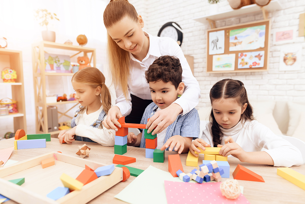 Kids playing with wooden cubes and teacher is helping them.